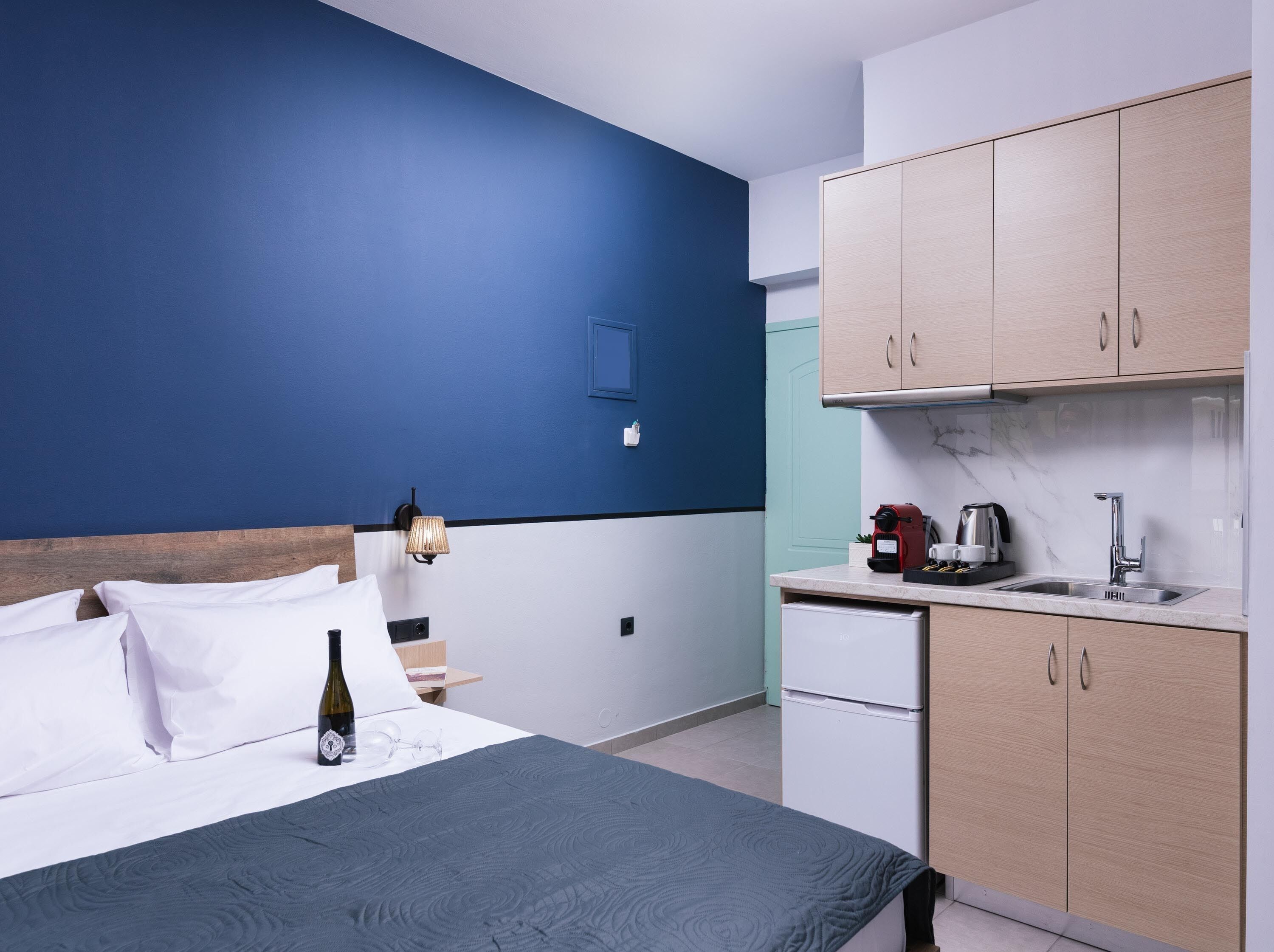 Une chambre d’hôtel comprenant un lit, une table de chevet, une lampe, une petite kitchenette et un mur d’accent bleu.