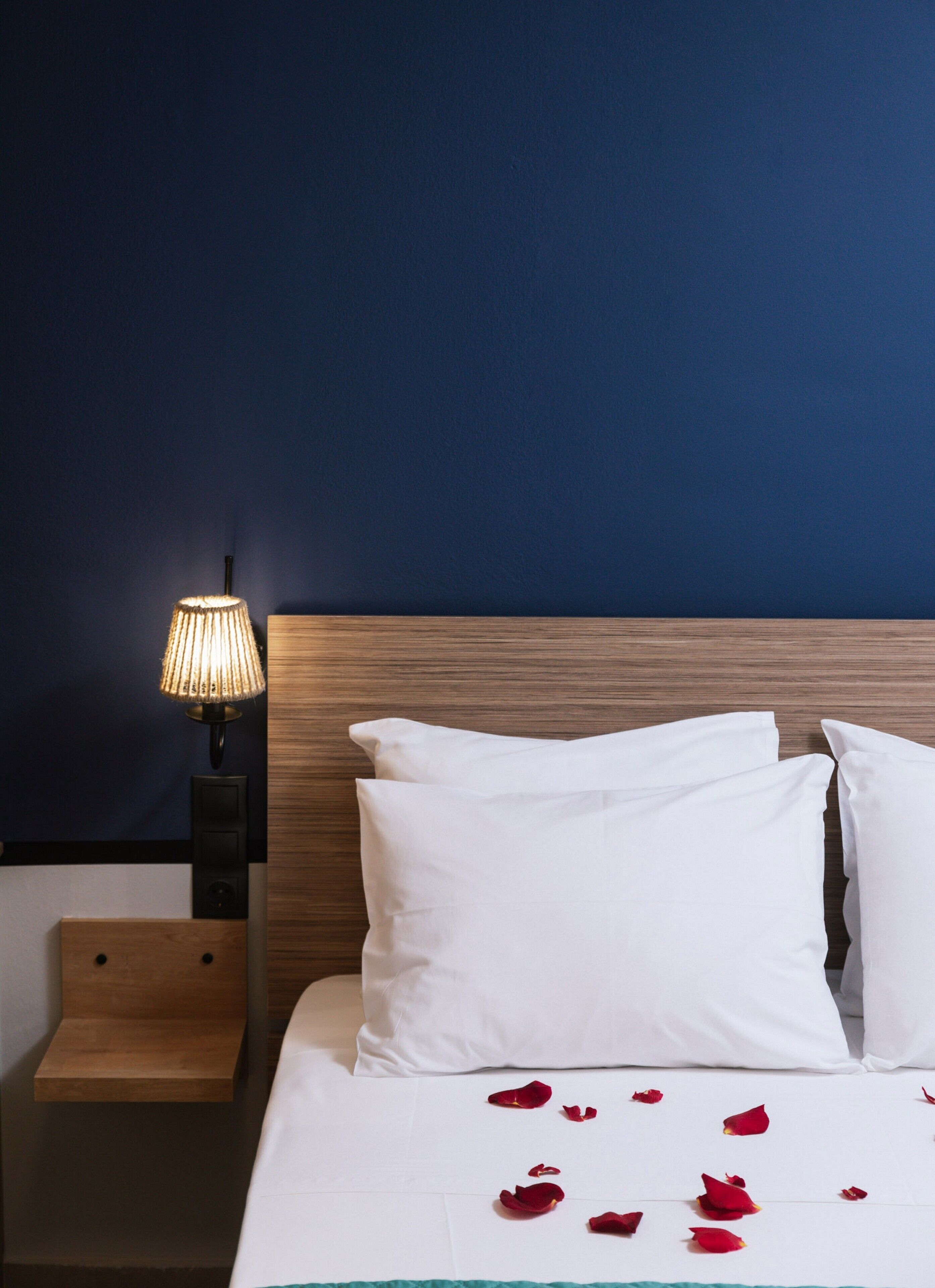 A neatly made bed with white bedding and rose petals scattered on it, a wooden nightstand with a lamp, and a dark blue wall.
