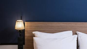 A neatly made bed with white bedding and rose petals scattered on it, a wooden nightstand with a lamp, and a dark blue wall.