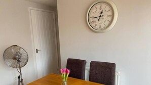 A dining area with a wooden table, two chairs, a vase with flowers, a wall clock, and a standing fan. - Cosy Homely Apartment Close to Lincoln City Centre (Lincoln)