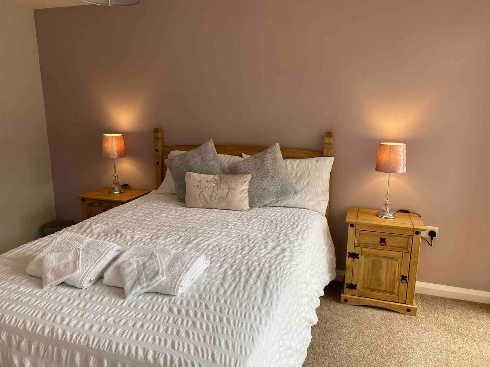 A neatly made bed with a white quilt, pillows, and a folded towel. There are two bedside tables with lamps and a wooden nightstand with a drawer.