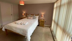 A neatly made bed with white linens and pillows, two bedside lamps, a wooden nightstand, and a large window with sheer curtains. - Cosy Homely Apartment Close to Lincoln City Centre (Lincoln)