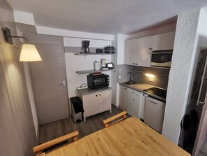 Uma cozinha compacta com micro-ondas, torradeira e cafeteira na bancada, uma pequena lixeira e uma mesa de madeira com bancos.