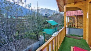 Un balcón de madera con vistas a montañas, un pequeño jardín y una estructura con techo rojo.