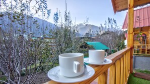 Una terraza de madera con dos tazas blancas sobre platillos, con vista a un jardín con árboles y una montaña al fondo.