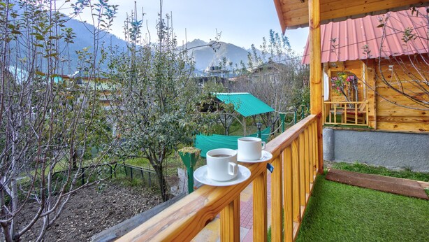 Un balcón de madera con dos tazas blancas sobre platillos, un paraguas verde y una vista de montañas al fondo.