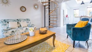 A spacious living area with a wooden coffee table, a blue sofa with tropical print, a blue armchair, and a spiral staircase leading to an upper floor. - Cozy Seaside Cottage with Terrace, Private Garden & Electric Car Charging (Vicq-sur-Mer)