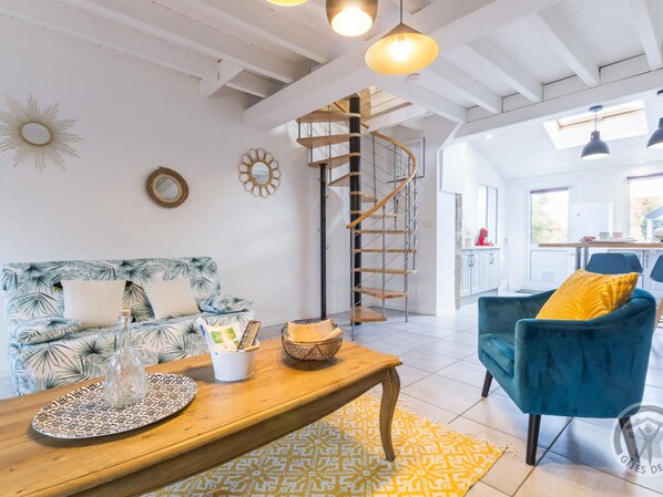 A spacious living area with a wooden coffee table, a blue sofa with tropical print, a blue armchair, and a spiral staircase leading to an upper floor.