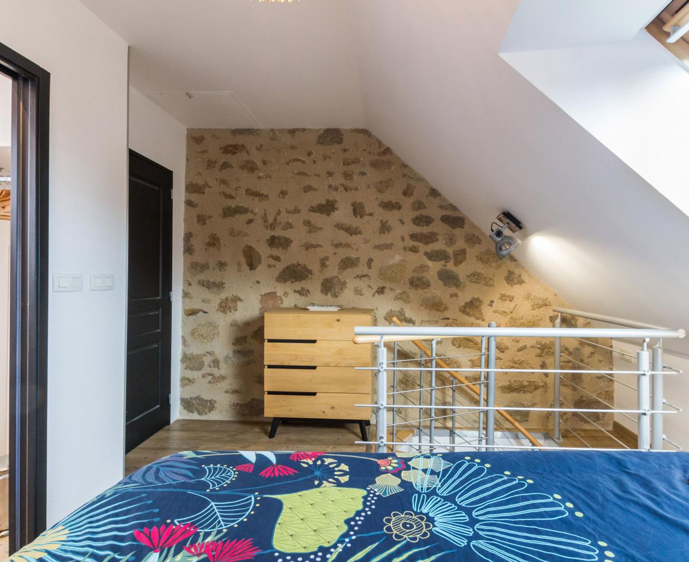 Ein Schlafzimmer mit schrägem Dach, einem hölzernen Schminktisch, einem Metallgeländer und einer gemusterten Bettdecke.