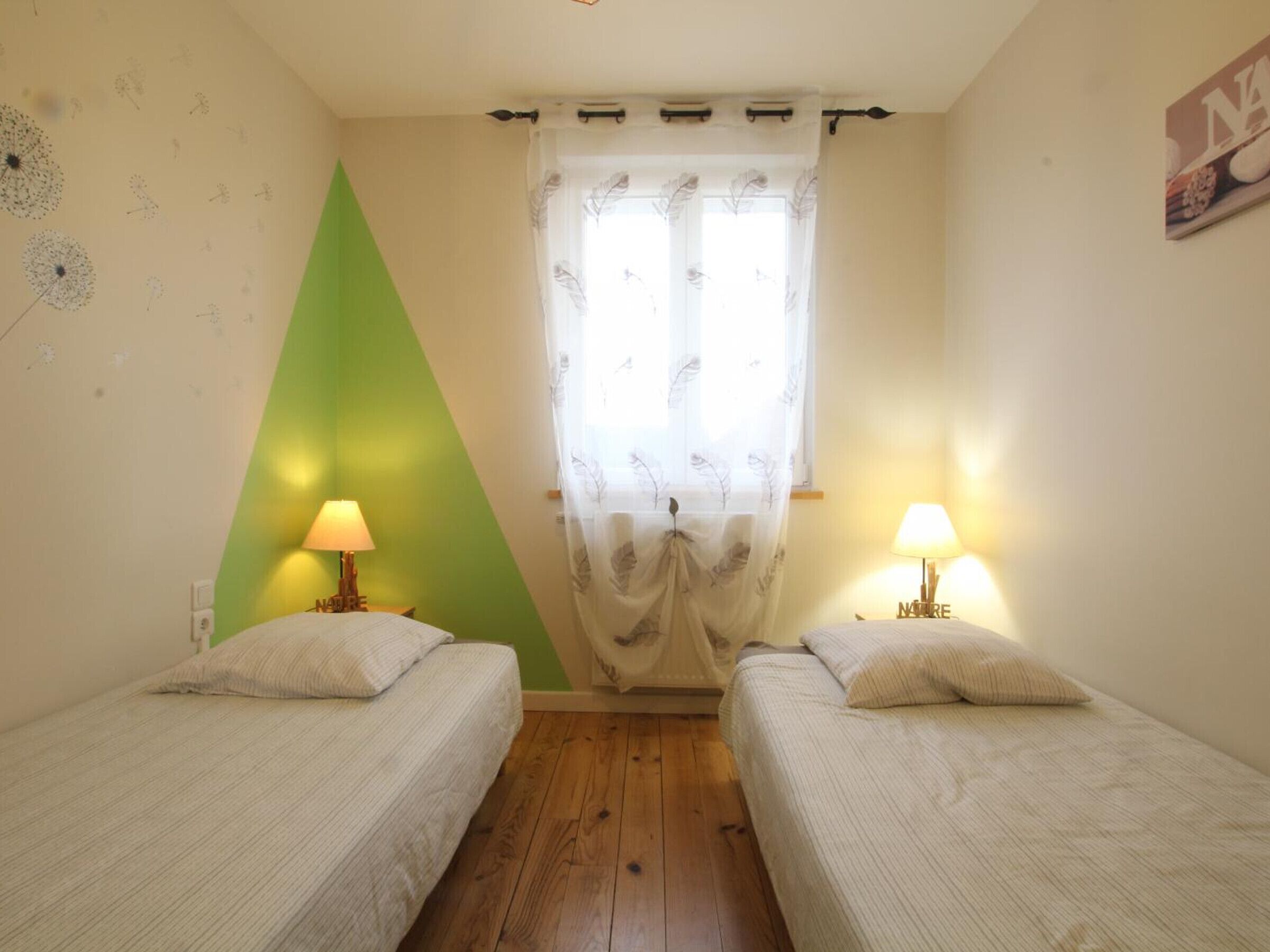 A room with two single beds, a green accent wall, a window with sheer curtains, and two bedside lamps.