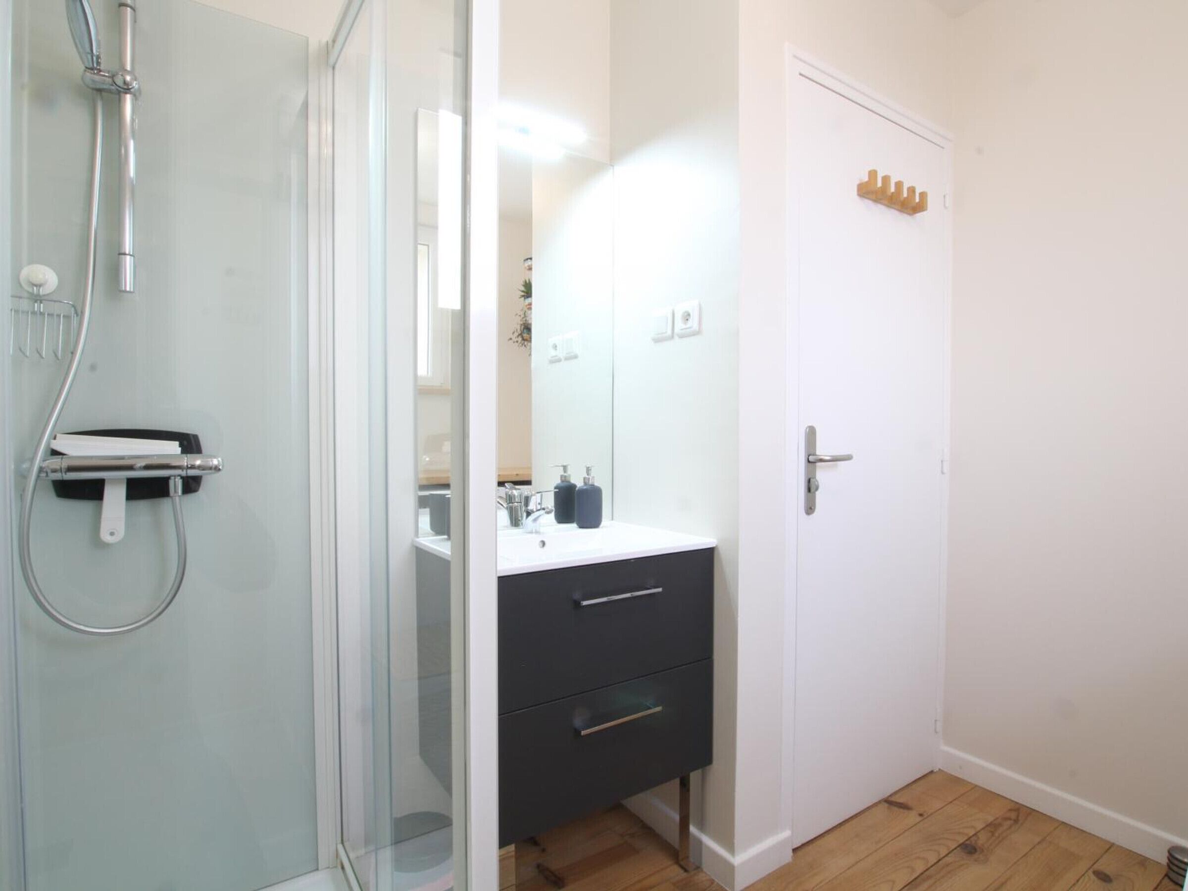 Un baño con cabina de ducha de vidrio, un lavabo blanco con mueble negro y una puerta blanca con toallero.