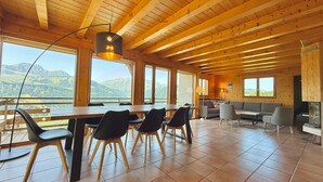 Una habitación amplia con techo de madera, grandes ventanas que ofrecen vistas a la montaña, una zona de comedor con sillas y una mesa, y un área de estar con sofás y televisor.