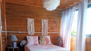 A wooden cabin bedroom with a bed, two nightstands, a hanging macrame decoration, and a large window with curtains. - L'ECRIN DUBUISSON Spacious villa in lush garden with whirlpool bath (Saint-Leu)