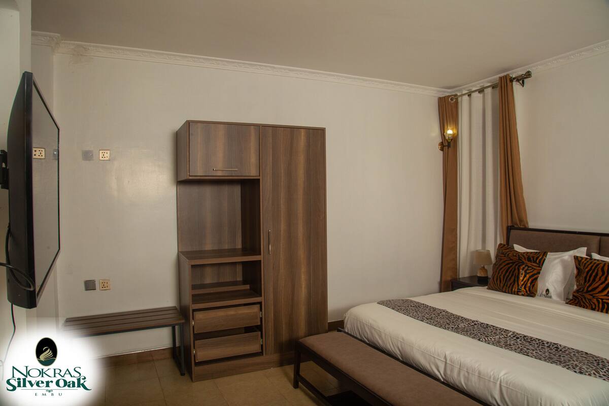 Quarto com cama, guarda-roupa de madeira, um banco e televisão.