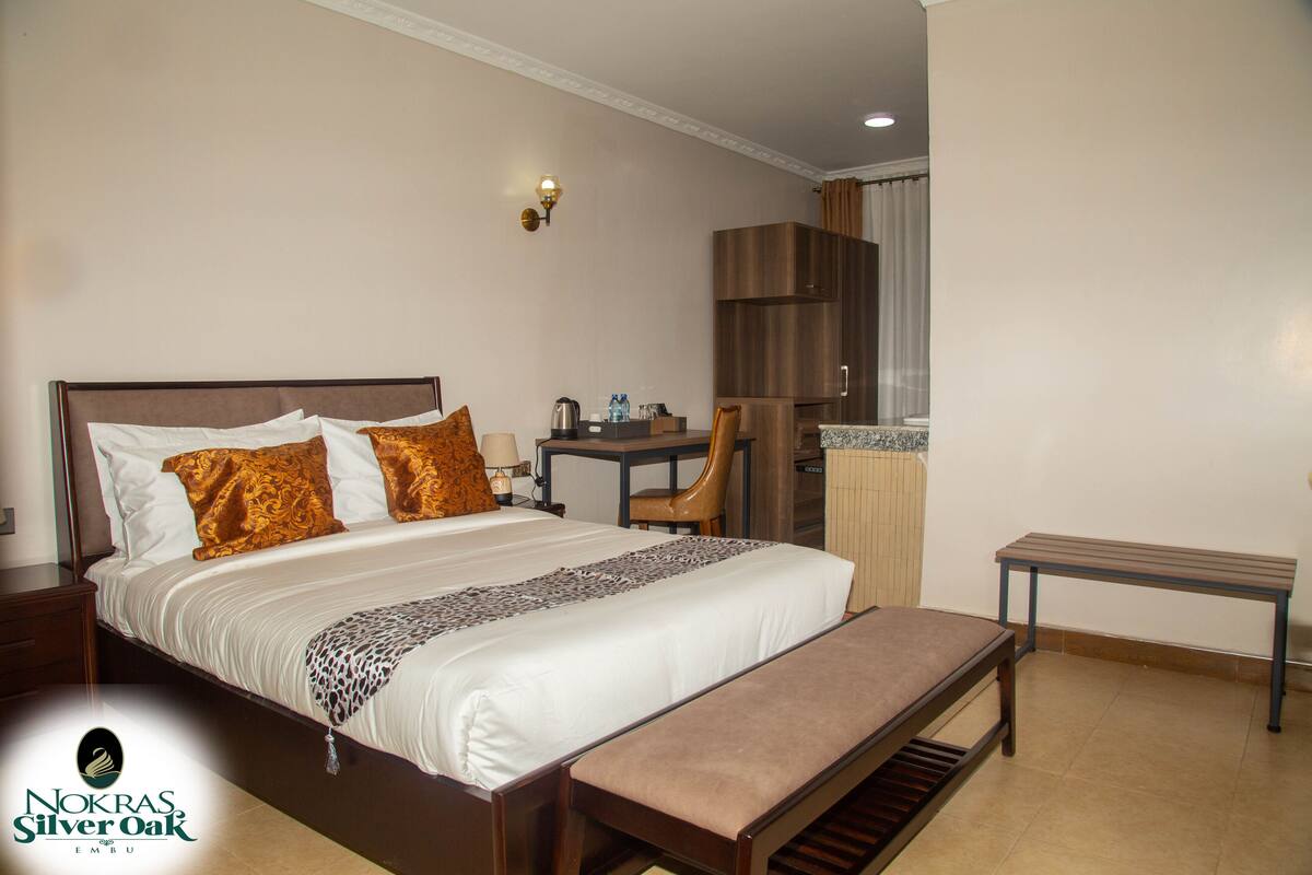 Quarto de hotel com uma cama grande, uma escrivaninha, uma cadeira, um banco e um minibar.