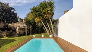 Una piscina rectangular con plataforma de madera, rodeada por un muro blanco y un área verde con palmeras.