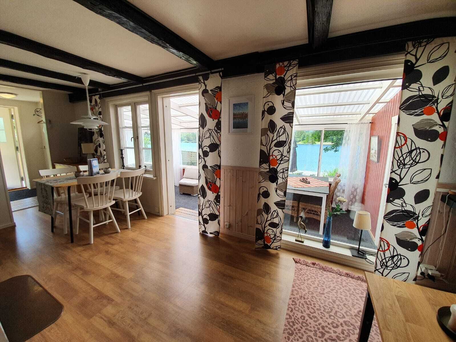A spacious room with a wooden floor, a dining area with a table and chairs, and a view of a lake through large windows.