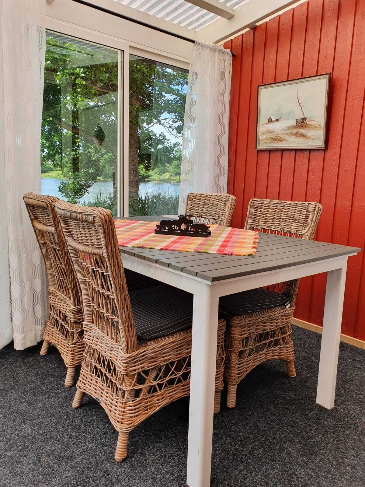 Ein Essbereich mit Korbstühlen, einem Tisch mit Platzdeckchen und Blick auf Grünpflanzen durch große Fenster.