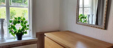 Une chambre d’angle avec une commode en bois, un miroir doté d’un cadre orné et une fenêtre donnant sur un paysage verdoyant.
