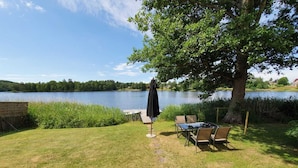 Un patio junto al lago con una mesa y sillas, una terraza de madera y un árbol grande que da sombra.
