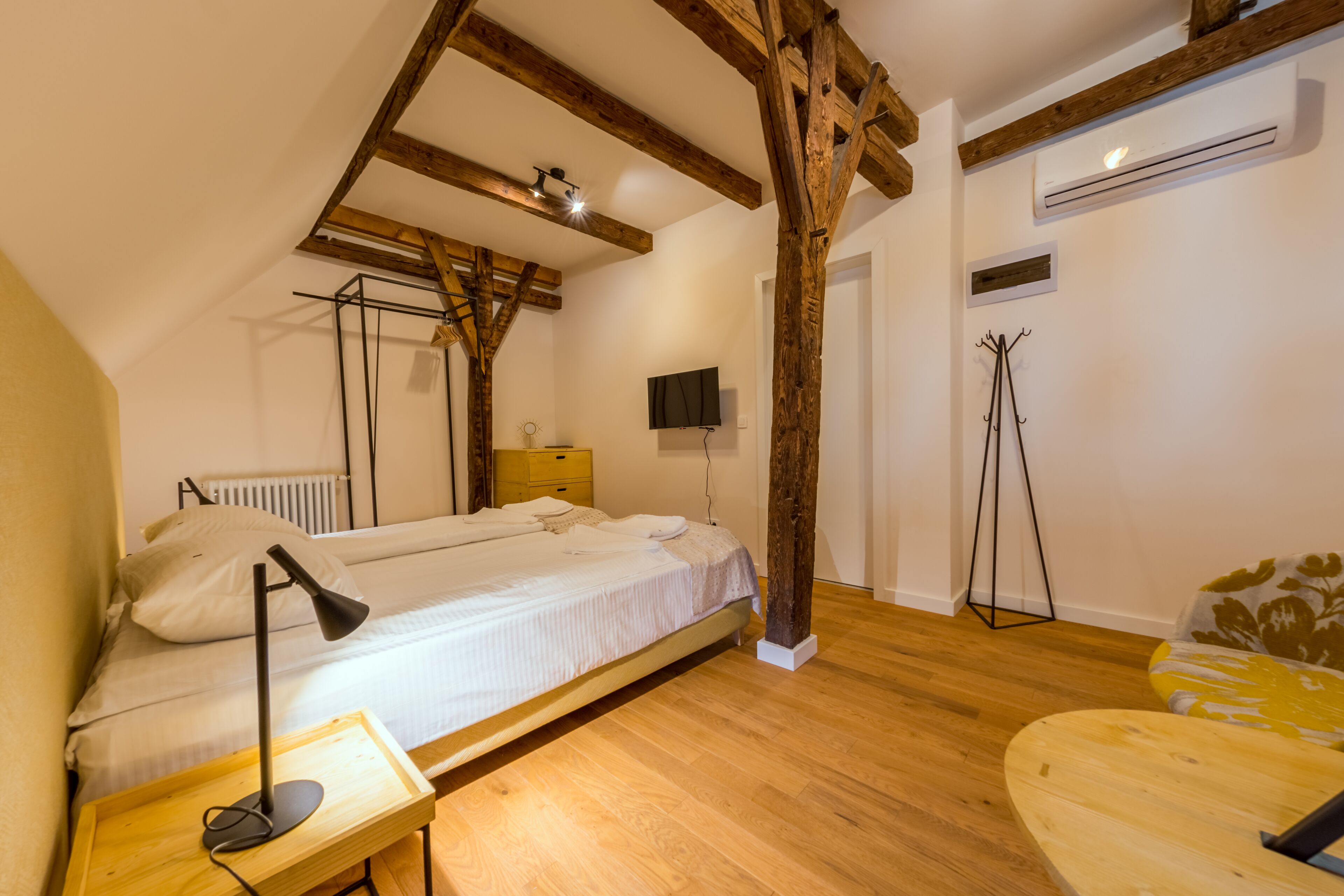 A bedroom with a wooden ceiling, a bed, a bedside table with a lamp, a TV, and a coat rack.