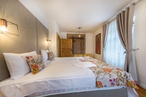 A neatly made bed with a floral bedspread and multiple pillows, a wooden wardrobe, and a large window with curtains.