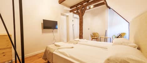 Une chambre avec un plafond en bois, un lit recouvert de draps blancs, un téléviseur fixé au mur et une chaise agrémentée d’un coussin à motifs floraux.