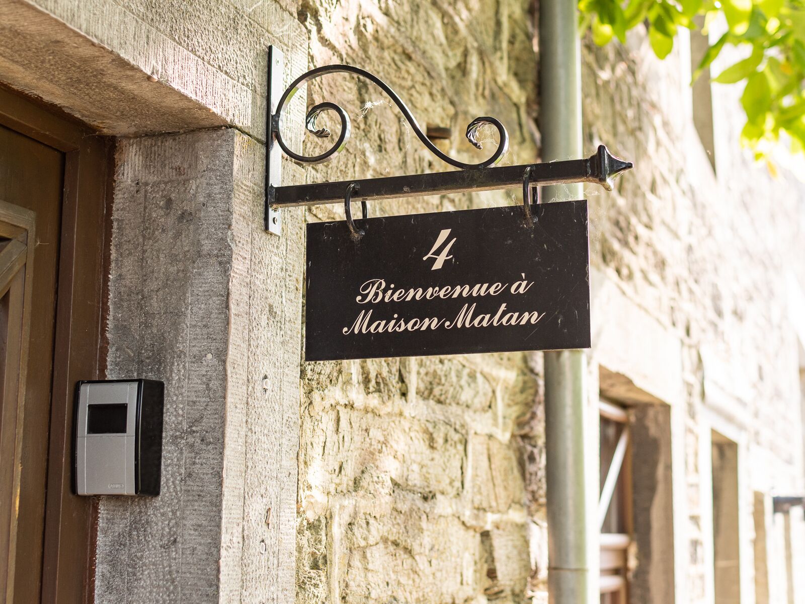 A black sign with white lettering on a metal bracket, reading "4 Bienvenue à Maison Matan" against a stone wall background.