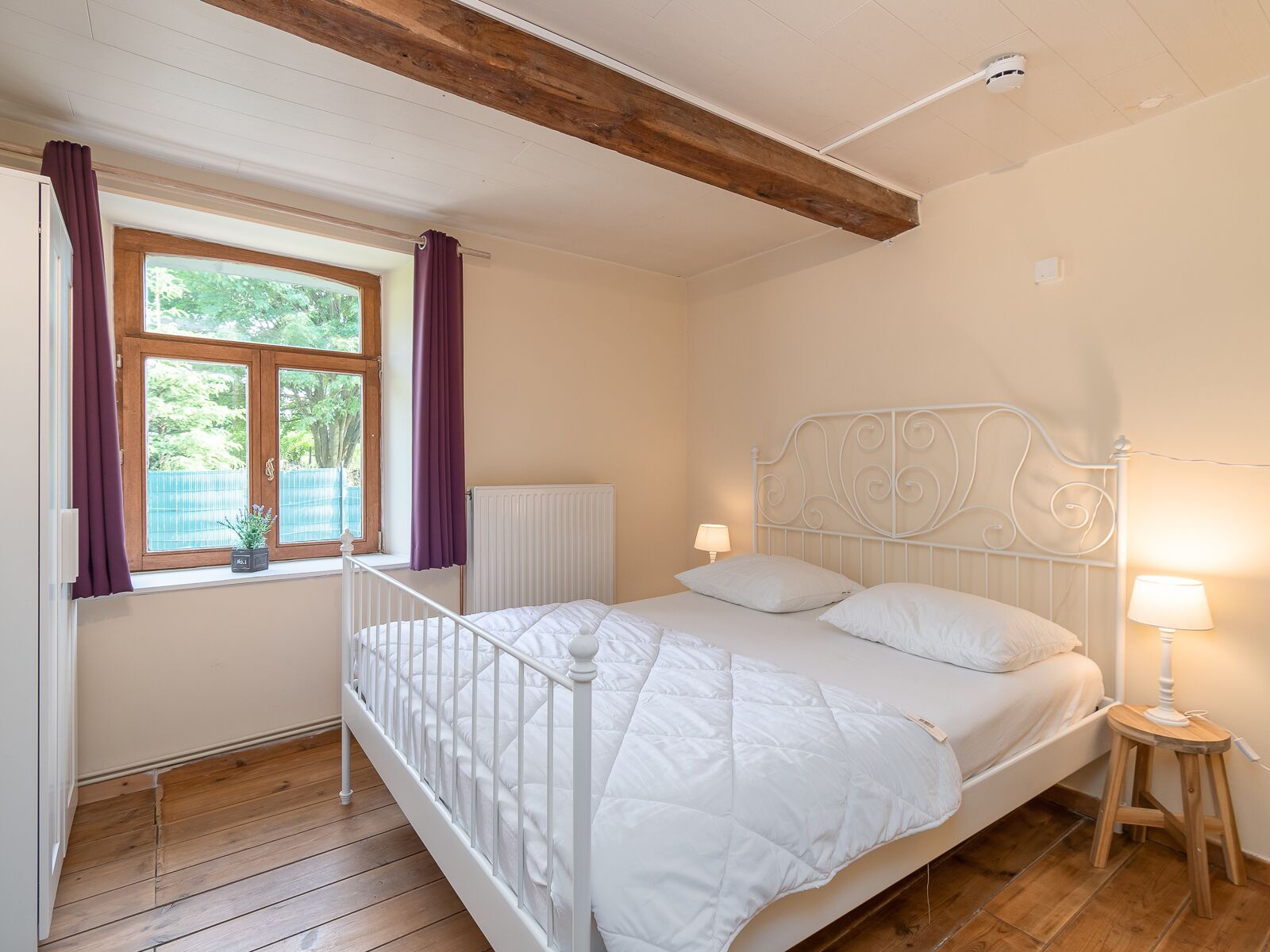 A bedroom with a white bed, wooden floor, a window with purple curtains, and a white radiator.