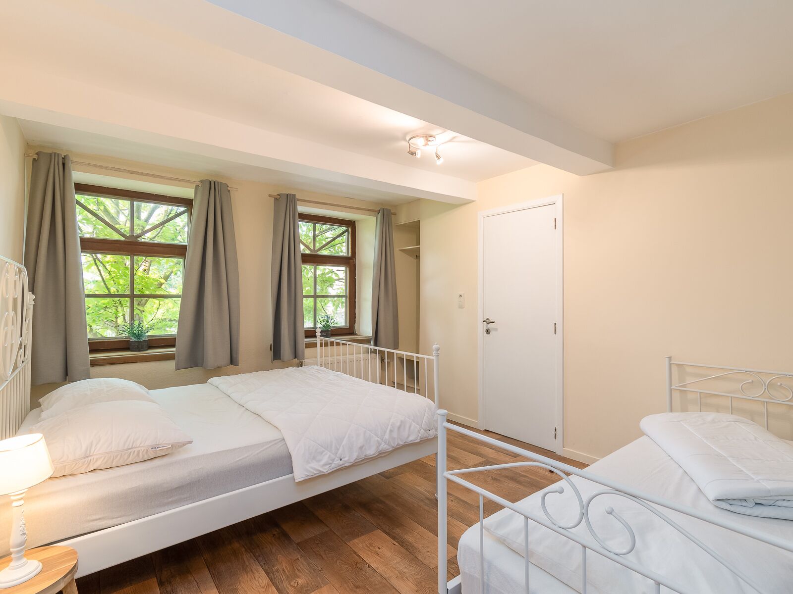 A bedroom with two beds, a window with curtains, a door, and a bedside table with a lamp.