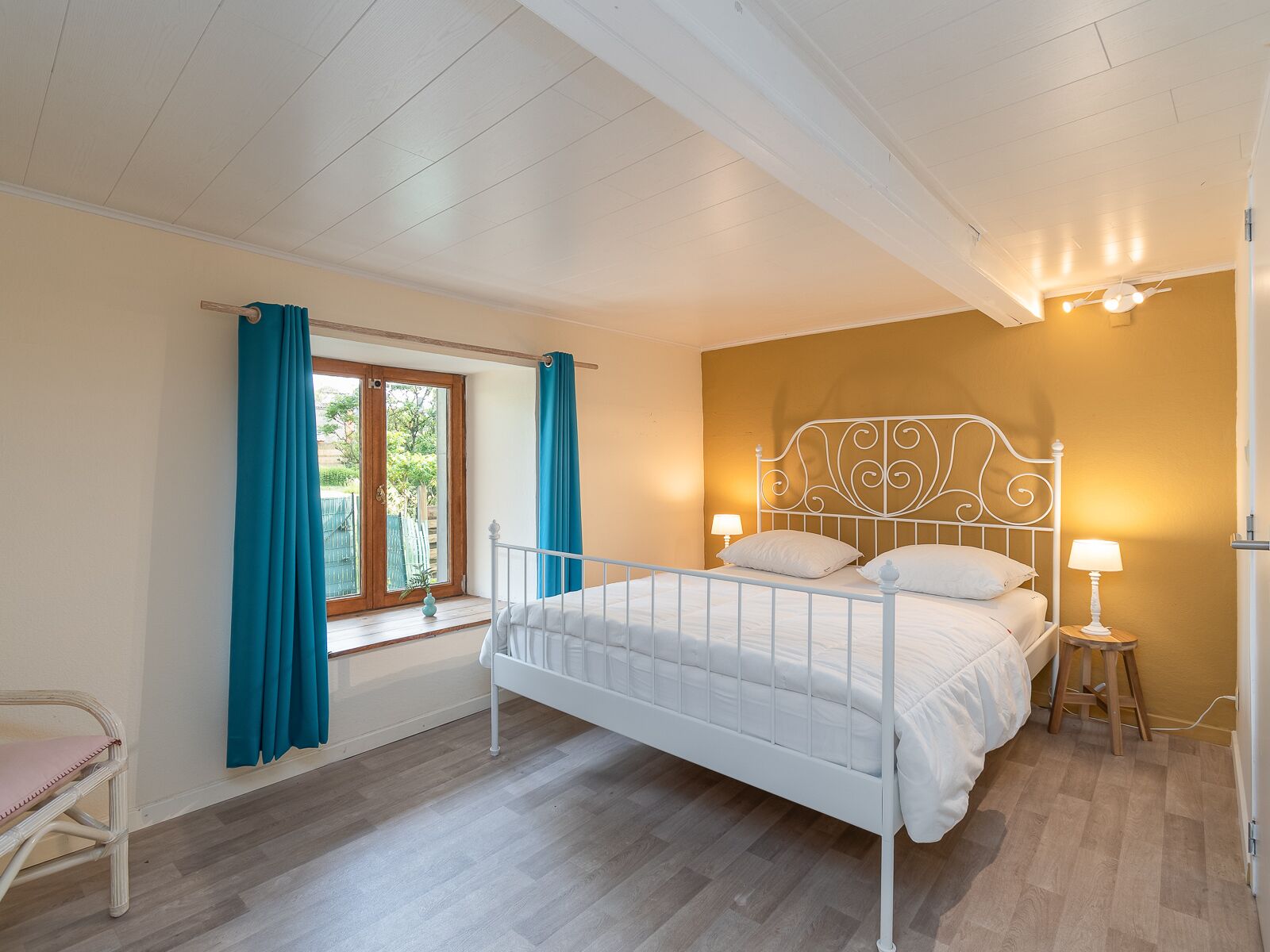 Ein Schlafzimmer mit einem weißen Metallbett, einem Fenster mit türkisblauen Vorhängen, einem Holzstuhl und einer Wandleuchte.