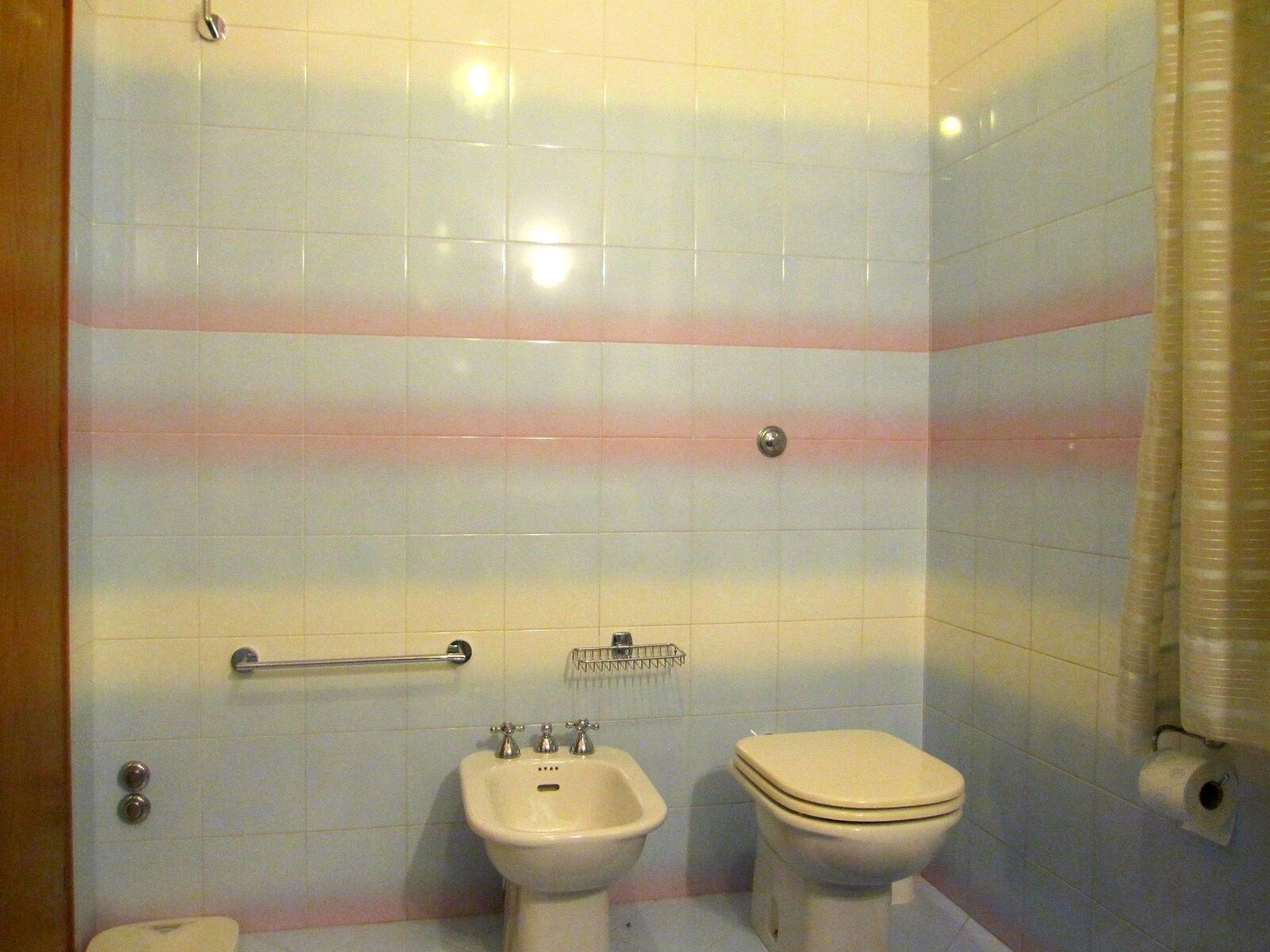 A bathroom with white tiles, a bidet, a toilet, a towel rack, and a hand dryer.