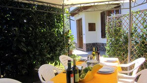 An outdoor dining area with a table set for four, surrounded by white plastic chairs. There are wine bottles, a fruit bowl, and plates on the table.