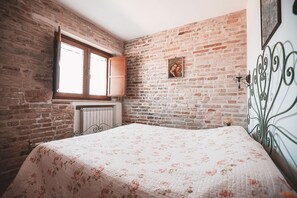 A bedroom with a brick wall, a window, a radiator, a bed with a floral bedspread, and a decorative metal headboard. - Apartment with a pool in the Tour des Palmiers with ocean views—5 minutes from the beach (Marche)