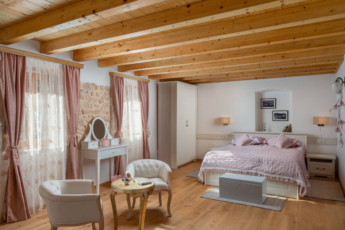 A bedroom with a wooden ceiling, a bed with pink bedding, a white dresser, a vanity with a mirror, and a chair.