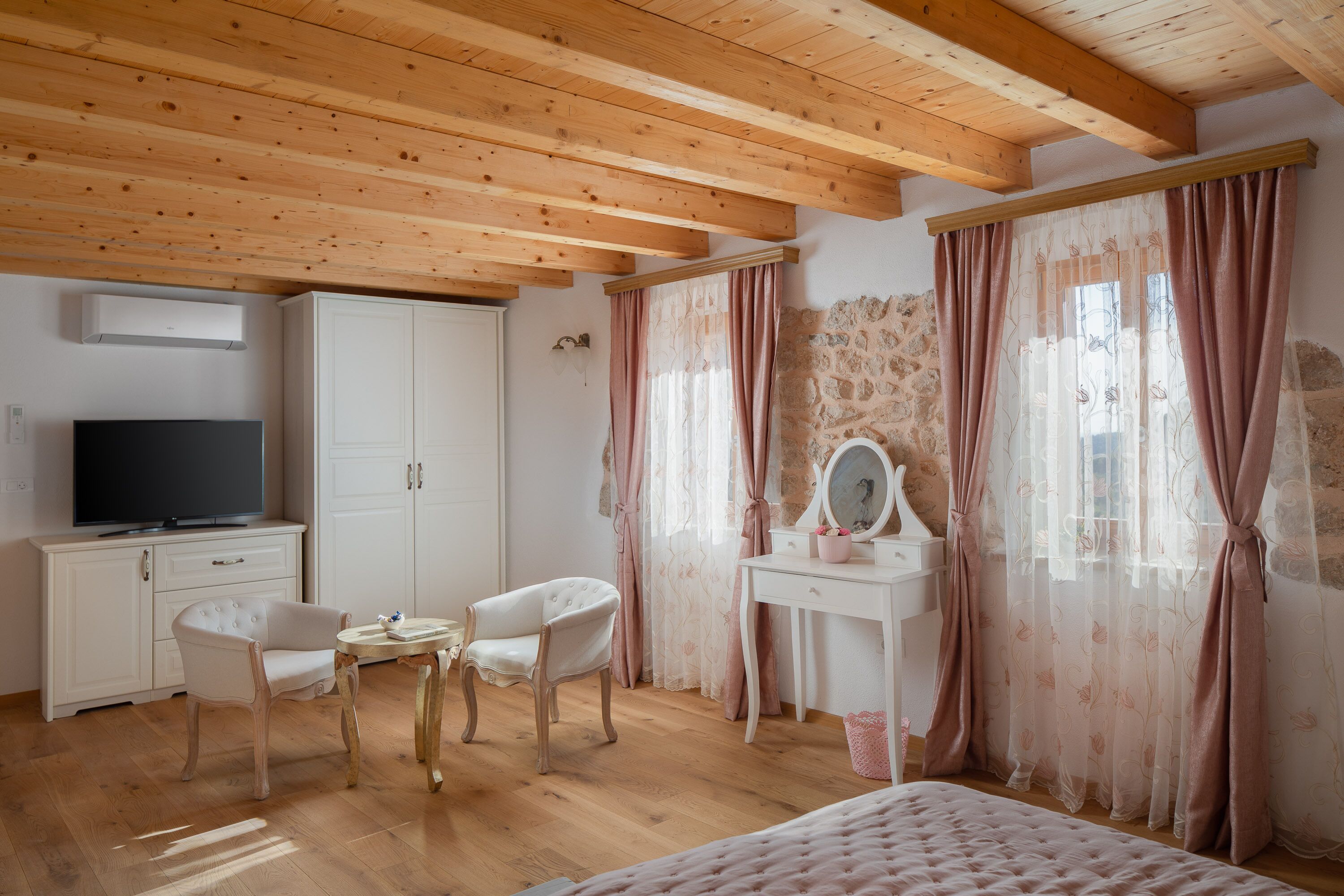 A bedroom with wooden beams, a TV, a wardrobe, a vanity with a mirror, and a bed.