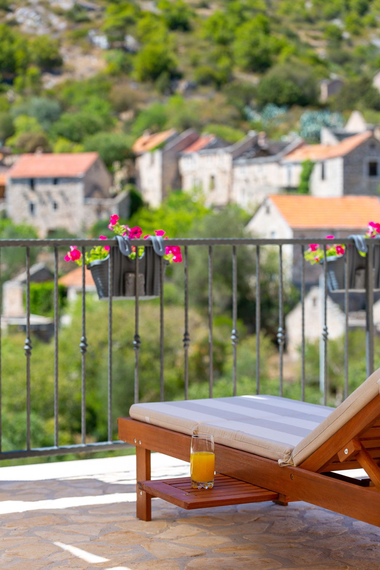 A wooden lounge chair with a cushion and a glass of orange juice on a stone-tiled patio.