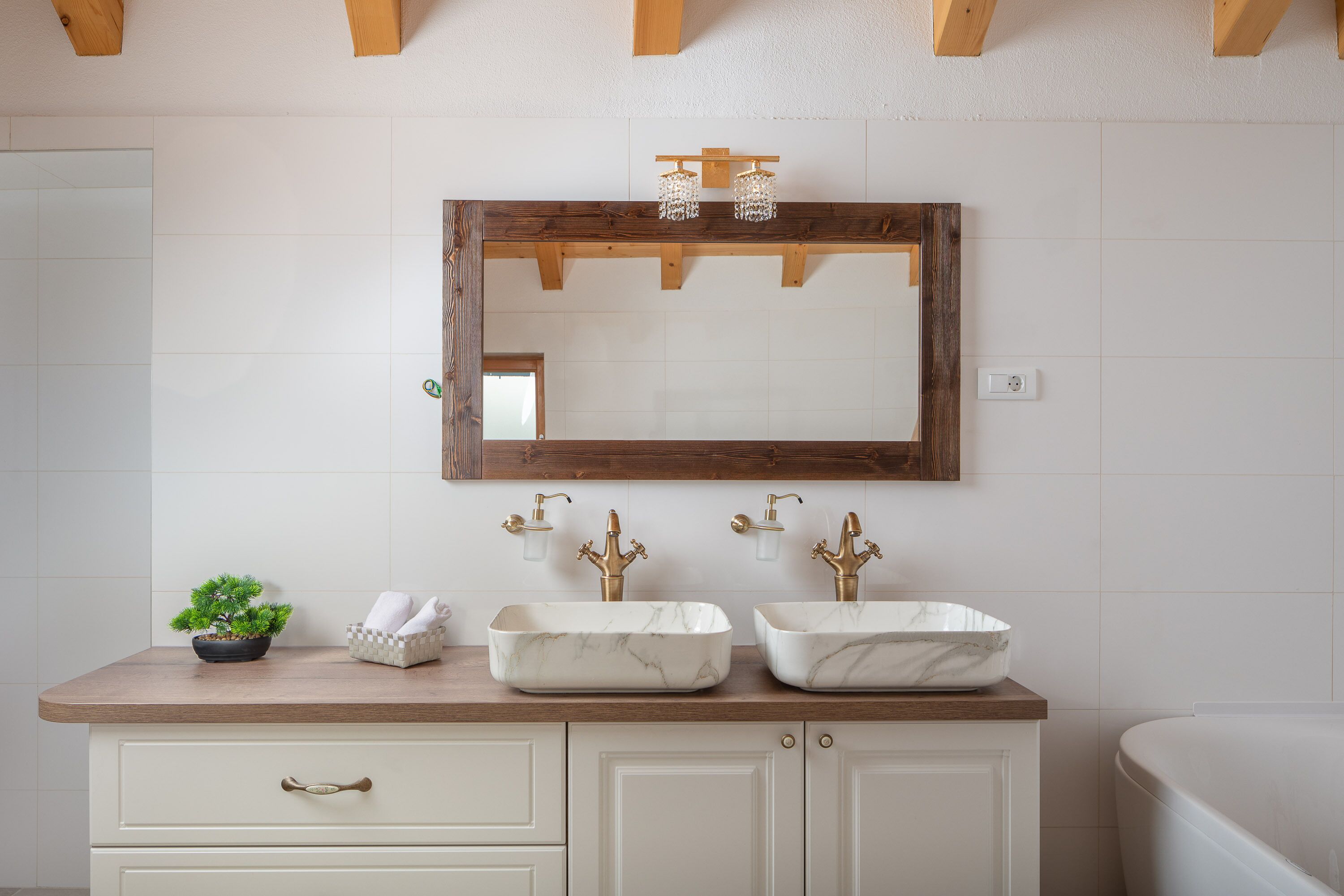 A bathroom with a wooden vanity, two marble sinks, a small potted plant, and a mirror with a wooden frame.