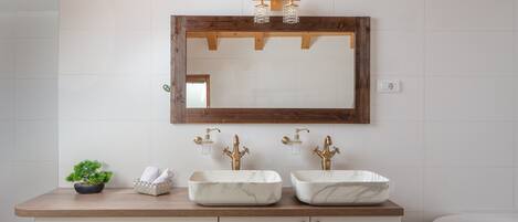 A bathroom with a wooden vanity, two marble sinks, a small potted plant, and a mirror with a wooden frame.