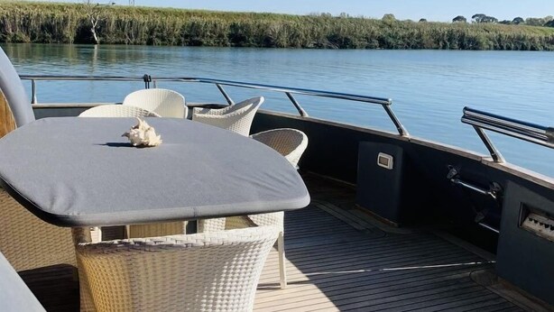 Cubierta de barco con una mesa y sillas, barandilla y vista a una masa de agua tranquila con vegetación en la orilla.