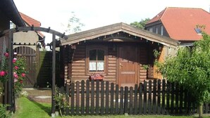 Een houten vakantiehuisje met een picket hek, een houten poort en een kleine tuin met roze bloemen.