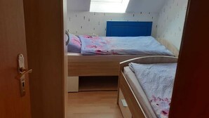 Een klein appartement met één kamer, een schuine kap en dakraam. Er is een bed met een blauwe hoofdeinde en een raam met jaloezieën.
