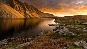Un lago sereno con montañas al fondo, rodeado de terreno rocoso y vegetación escasa.