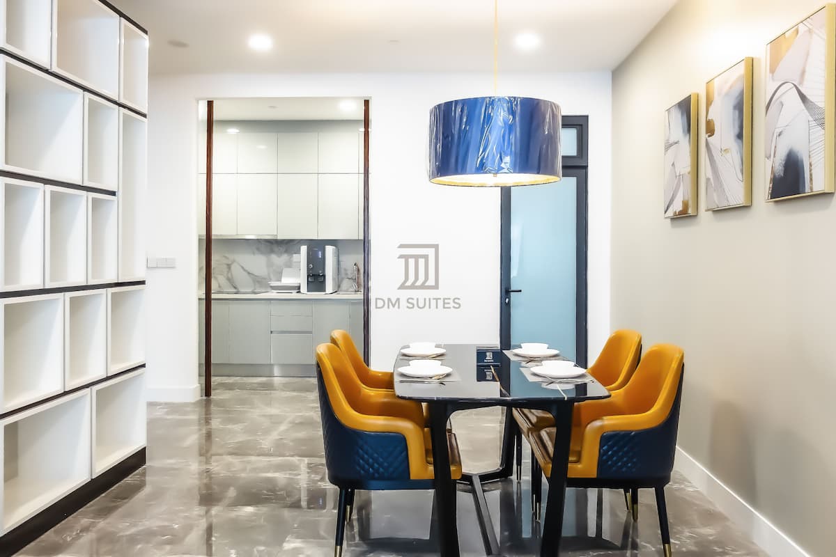 A modern dining area with a table set for four, a blue pendant light, and a kitchen in the background.