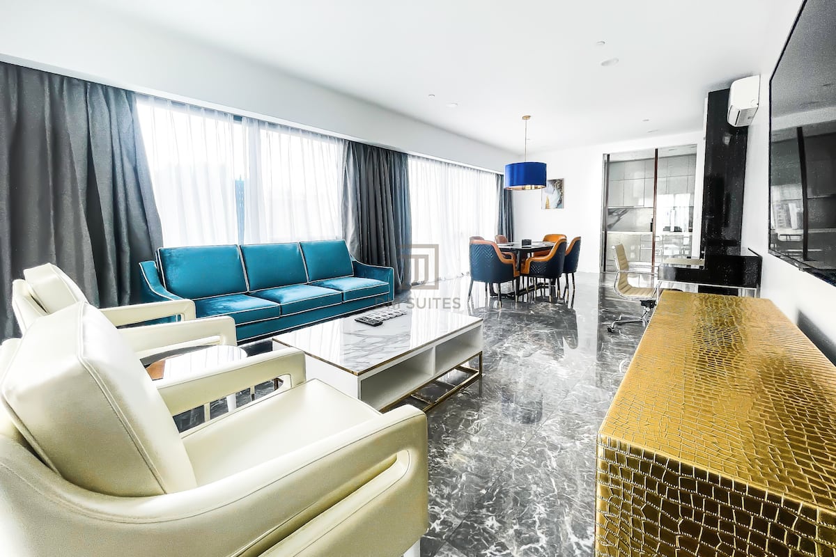 A modern living room with a blue sofa, a white armchair, and a glass coffee table. There is a dining area with a table and chairs, and a kitchen area with a black countertop and a large window.