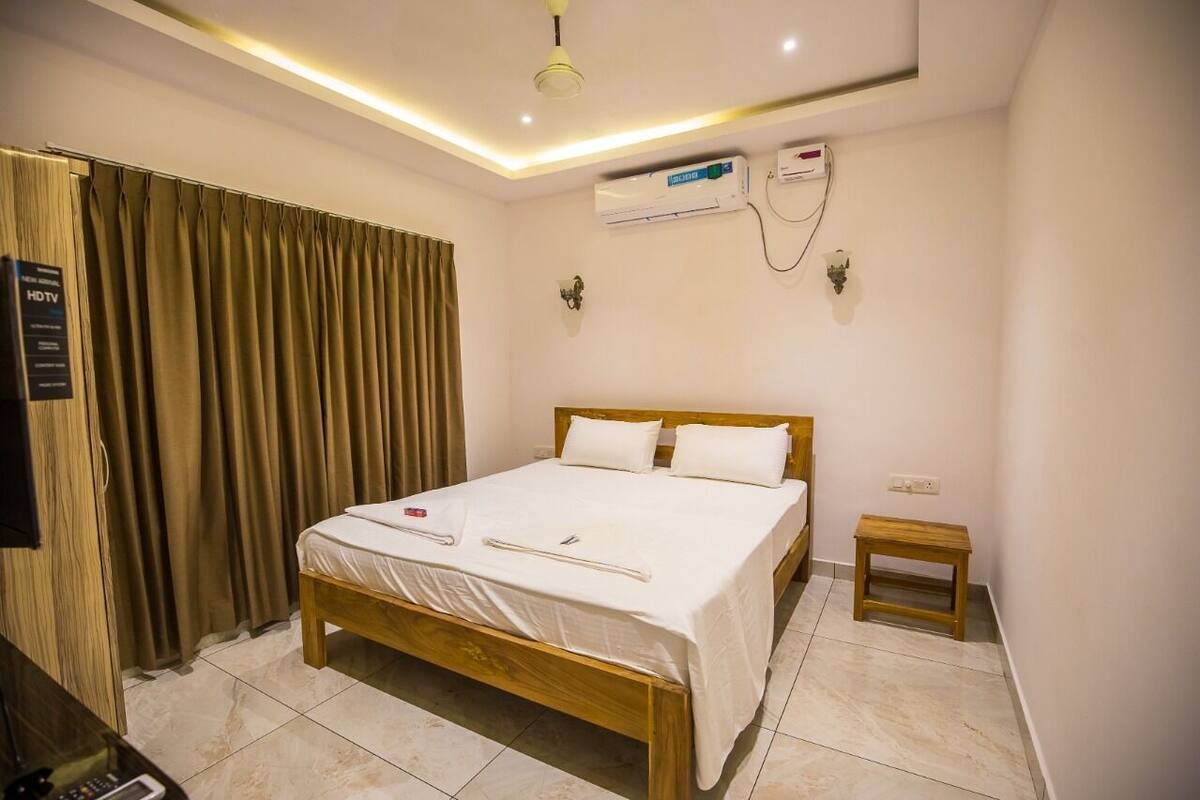 A hotel room with a wooden bed, white bedding, a small wooden stool, an air conditioning unit, and a ceiling light.