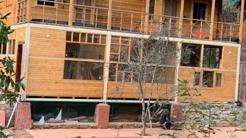 A two-story wooden cabin with a balcony, surrounded by a stone wall and vegetation.