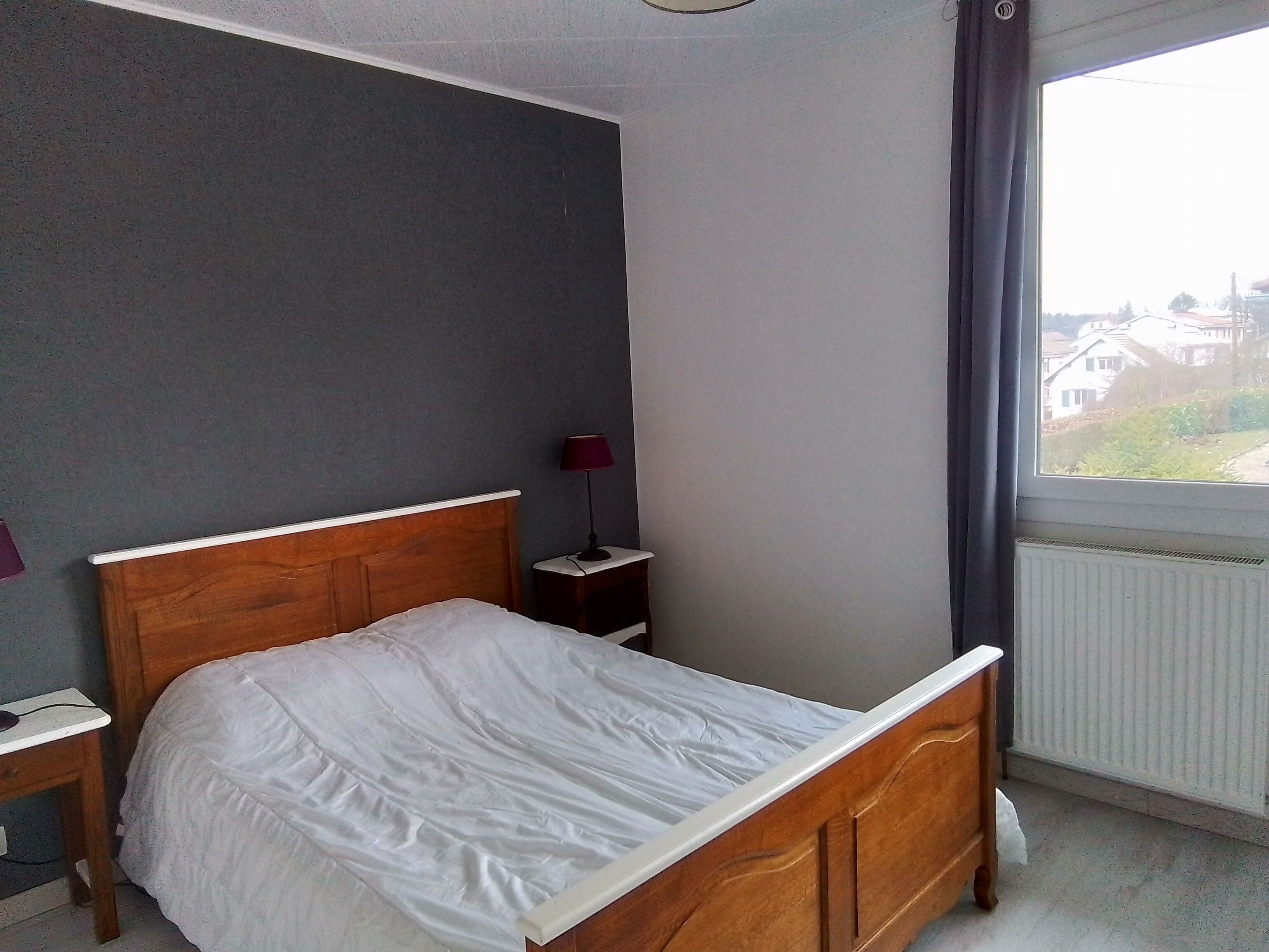 Un dormitorio con cama de madera, mesita de noche, lámpara y ventana con vista a edificios.
