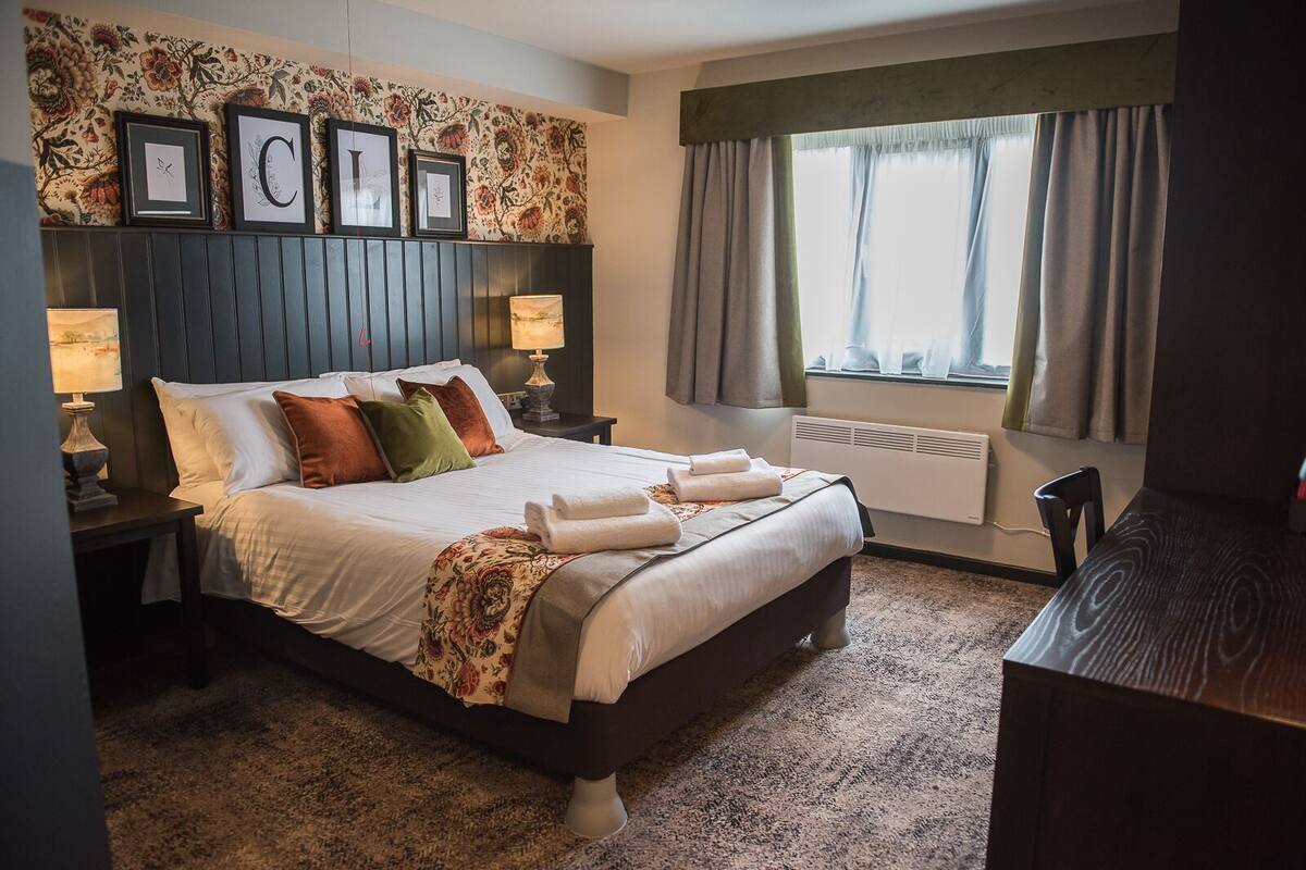 A bedroom with a bed, nightstands, a desk, a radiator, and a window with curtains.
