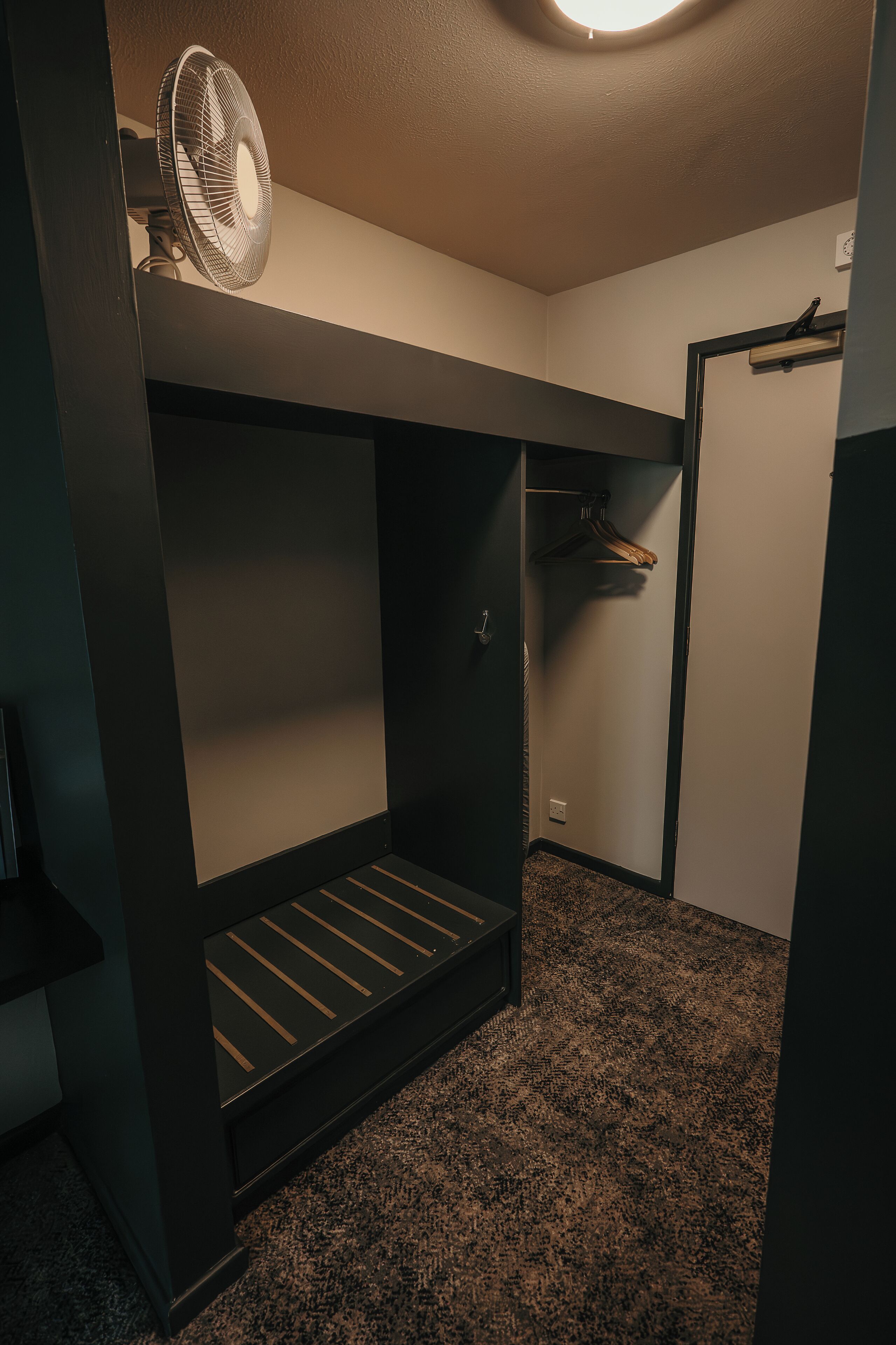 Um quarto pequeno e com pouca luz, com uma cama de solteiro, um ventilador de teto e um cabideiro.